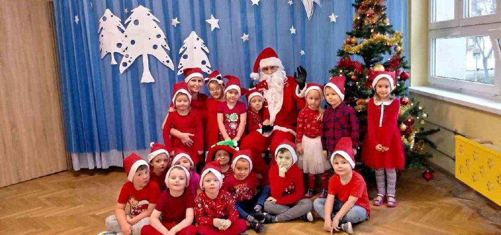 Dzieci w czerwonych strojach pozują razem z Mikołajem na tle choinki. Z tyłu niebieskie tło z białymi choinkami i białymi gwiazdkami.